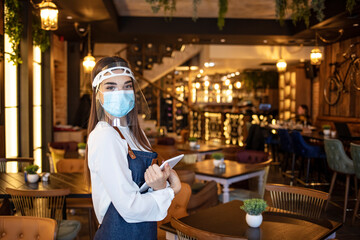 Female waitress wearing protective face mask and visor while holding touchpad and reopening...
