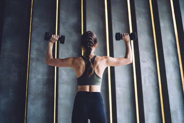 Rear view of female in trendy activewear with muscular figure lifting weight during workout, back view of woman slimming and reaching fitness goals on workout for arms and back muscles in gym