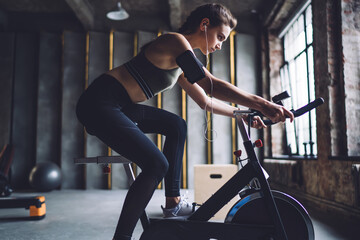 Fototapeta na wymiar Serious caucasian female in activewear sitting on exercising bike ready for reaching good results in cardiom determined woman listening online radio via sound accessory slimming and losing weight