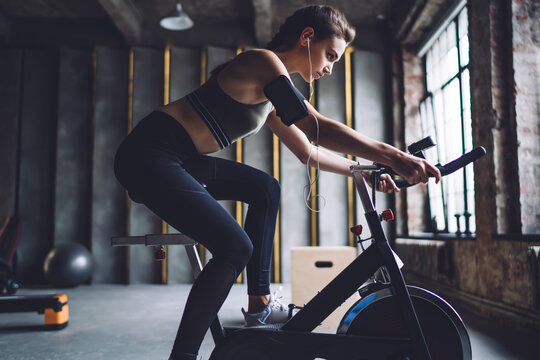 Serious caucasian female in activewear sitting on exercising bike ready for reaching good results in cardiom determined woman listening online radio via sound accessory slimming and losing weight
