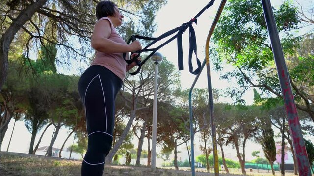 A brunette Portuguese woman, middle aged, doing a straps squat, in a park on a sunny morning.
