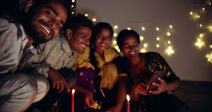 Slow-motion medium shot of friends, males and females, take selfie photo mobile smart phone camera technology Digital device during Diwali celebration decoration at home 