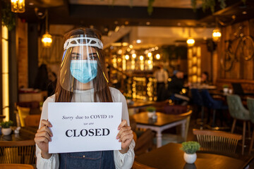 Female barista holding closed sign when standing at coffeeshop entrance. Female owner holding closed sign at indoor restaurant due to coronavirus pandemic.