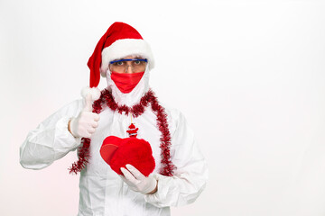 Fototapeta na wymiar Health worker wearing protective clothing. He wears a santa hat on his head. He has a red corona mask on his face. He is holding a gift box in the shape of a heart.