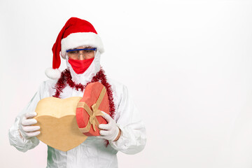 Health worker wearing protective clothing. He wears a santa hat on his head. He has a red corona mask on his face. He is holding a gift box in the shape of a heart.