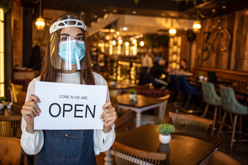 Young Caucasian female waitress wearing a face mask, hanging a sign that says Open onto a door of her cafe. Small business owner with face mask holding the sign for the reopening of the place 