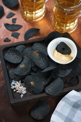 Close-up of black potato chips on a wooden serving tray and mugs of beer in the background, vertical shot