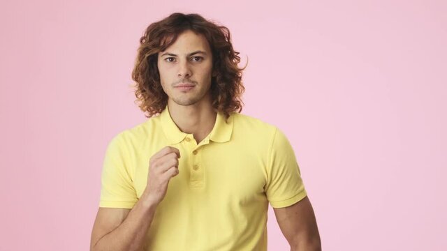 A focused curly young man is watching something then feeling disappointed standing isolated over a pink background in the studio