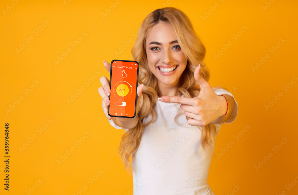 Poster young lady pointing at mobile phone with smarthome app on screen, regulating temperature over orange