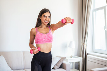 Fototapeta na wymiar Woman exercising with dumbbells at home. Sporty beautiful woman exercising at home to stay fit. Young woman exercising at home in a living room. Fitness, workout, healthy living and diet concept.