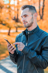 Youthful Guy Using Black Smartphone at the Beautiful Autumn Park. Handsome Young Man with Mobile Phone at Sunny Day - Medium Shot Portrait