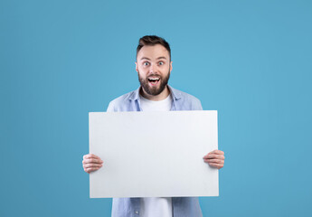 Excited young man holding blank poster with copy space on blue studio background