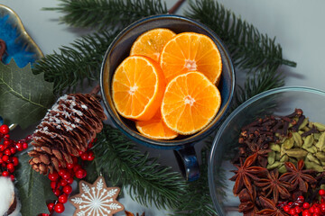 Fototapeta na wymiar Christmas items on the table. Pieces of tangerines in a cup, fir twigs and cones, winter spices, decorations in the form of ginger cookies, holly berries and more