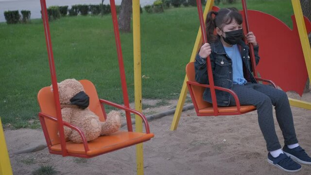 Thoughtful girl in mask. A nice pensive little girl sits on the swing by the teddy beath both in medical facial mask. A concept of lonely isolation on the playground.