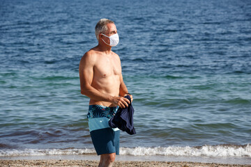 Uomo cinquantenne a petto nudo con mascherina protettiva sta in spiaggia di fronte al mare