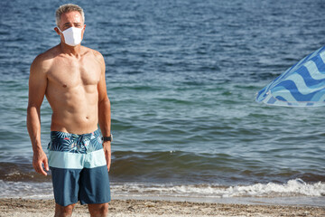 Uomo cinquantenne a petto nudo con mascherina protettiva sta in spiaggia in costume, isolato su sfondo mare
