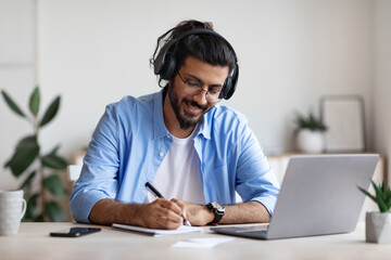 Eastern Guy Wearing Headphones Studying Online With Laptop At Home, Taking Notes