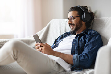 Home Leisure. Young Eastern Guy In Headphones Relaxing With Smartphone On Couch