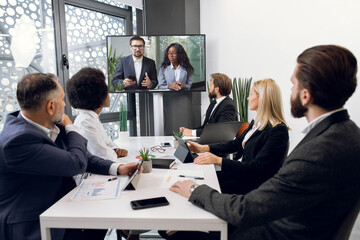 Focused multiracial business people in conference room, looking at a screen, having video conference with another team, African lady and Caucasian man, using smart video technology to communicate