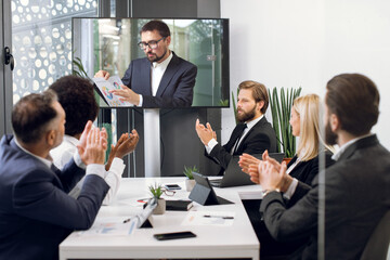 Group of five skillful multiethnical businesspeople clapping hands and looking at screen, having video conference in boardroom, with their male colleague explaining some financial charts
