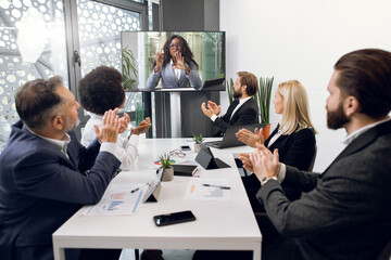 Videoconference, telemeeting, remote work. Multiethnic diverse business team, sitting around the table in office room, greeting their female African colleague, clapping hands and looking at tv screen