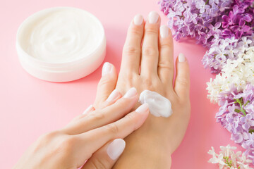 Obraz na płótnie Canvas Young perfect adult woman hands using white moisturizing cream. Care about nails and clean, soft, smooth body skin. Beautiful branches of fresh, colorful lilac flowers. Closeup. Point of view shot.