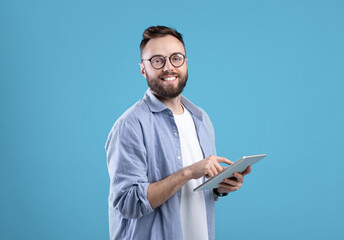 Naklejka na ściany i meble Positive young guy in casual wear using tablet computer over blue studio background