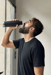 Man in black sportswear drinking water by the window in a white gym interior, portrait