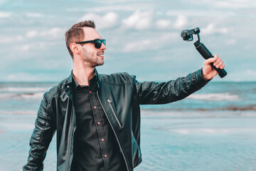 Portrait of Smiling Blogger in Sunglasses Making Selfie or Streaming Video at the Beach Using Action Camera with Gimbal Camera Stabilizer. Man in Black Clothes Making Photos