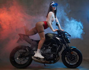 Fototapeta na wymiar Sexy graceful and slim woman in short clothing with cap poses with on urban custom motorbike in smokey shiny studio.