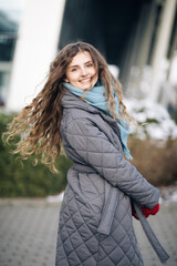 Charming young curly woman with a magnificent brown hair, big brown eyes and stylish look turns to camera and smiles. Attractive young lady is rushing in the winter city-center.