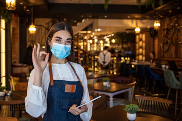 Fototapeta na wymiar Positive thinking during the crisis caused by the coronavirus, young woman waitress in uniform, wearing gloves and protective mask at work in the restaurant, makes the ok gesture with the hand