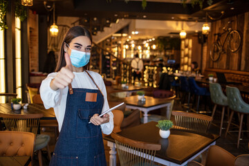 Shot of a handsome young woman welcoming customers on the door of the store. Come on in, we're open. Portrait of a Caucasian woman with a jeans apron, wearing a face mask showing thumb up.