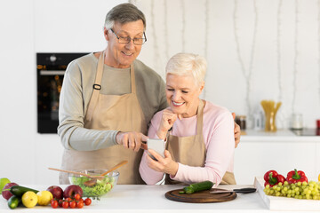 Cheerful Senior Spouses Using Cellphone Cooking Dinner In Kitchen