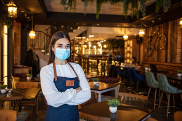 Fototapeta na wymiar Portrait of a young Caucasian female waitress wearing an apron, face mask and gloves, standing at the entrance of a drink establishment and holding a digital tablet computer