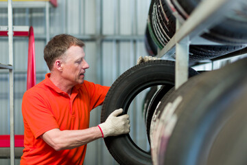 Car service. Male worker checking new tire wheel on shelves shelf at wheel store, wearing red...