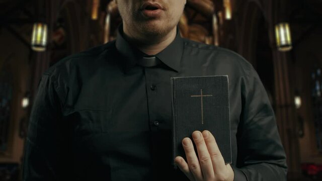 Closeup portrait of a young catholic praying priest on the background of the church.Priest holding is dressing in black and reading New Testament book. Low-key image.