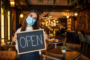 Portrait shot of beautiful woman hanging up an open sign on her store window. Welcome back to our store. Young Caucasian female waitress wearing a face mask, hanging a sign that says Open 
