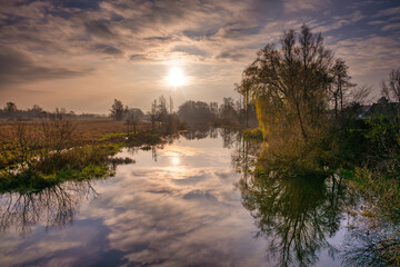 A backlit photo of tree and cloud reflections in a river. Beautiful sunset sky in the background. Picture from Scania county, southern Sweden