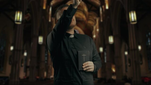 Closeup portrait of a young catholic praying priest on the background of the church.Priest holding is dressing in black, holding a crucifix,baptizing the room and reading New Testament book.