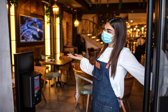 Beautiful Young Female Worker With Protective Mask On Face Working In Restaurant. Happy Female Cafe Owner Wearing Protective Face Mask While Standing At Entrance Door And Reopening After Coronavirus 