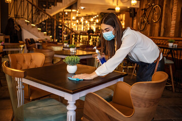 waitress disinfecting tables while wearing protective face mask due to coronavirus epidemic. Waitress wearing protective face mask while cleaning tables with disinfectant during COVID-19 epidemic.