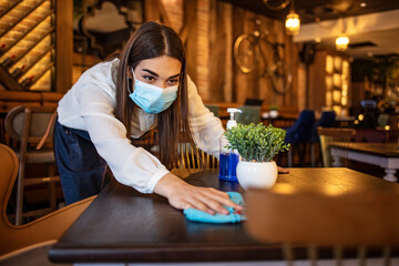 Young waitress disinfecting tables while wearing protective face mask due to coronavirus epidemic. Waitress wearing protective face mask while cleaning tables with disinfectant during COVID-19 