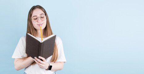 Portrait of thoughtful pensive girl having notebook and pencil in mouth. Isolated on blue background with copy space.