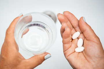 Female elderly hand with pills medicine tablets and glass of water against white sinks with fresh water. Senior woman taking pills at home, closeup. Drug therapy prescribed by a doctor