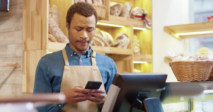 Close up of African American young handsome male worker in apron stands in bakery shop, texting and typing on smartphone. Man seller browsing online on cellphone. Business concept. Retail industry