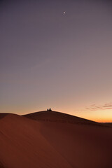 Fototapeta na wymiar Die Silhouette von zwei Personen auf einem Sandberg in der Wüste, der den Sonnenuntergang und einen Halbmond am Himmel beobachtet