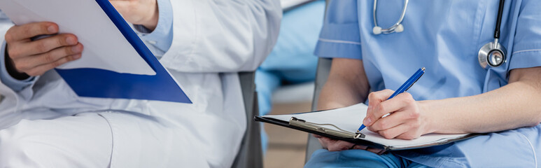 Nurse writing on clipboard while sitting near doctor during meeting in hospital on blurred background, banner