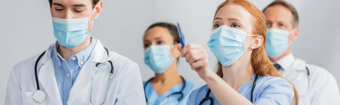 Redhead Nurse In Medical Mask Pointing With Pen During Meeting With Blurred Multiethnic Colleagues On Background, Banner