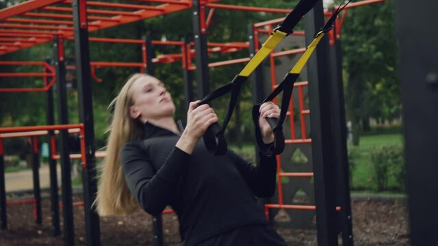 Woman in sports. Young athletic female in sportswear trains with trx fitness straps on the sports ground. Medium shot.